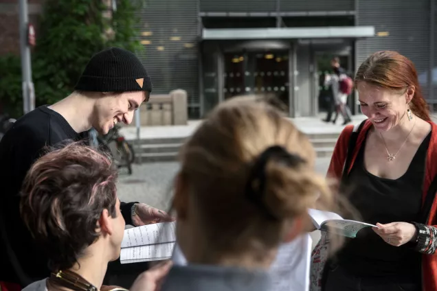 Four students laughing while holding and looking at books together in front of the LUX building 