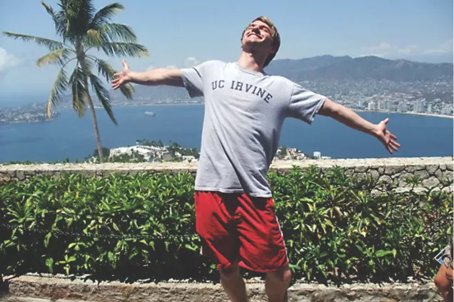 A student with an Irvine t-shirt smiles and stretches out their arms in front of the sea and a skyline. Photo. 